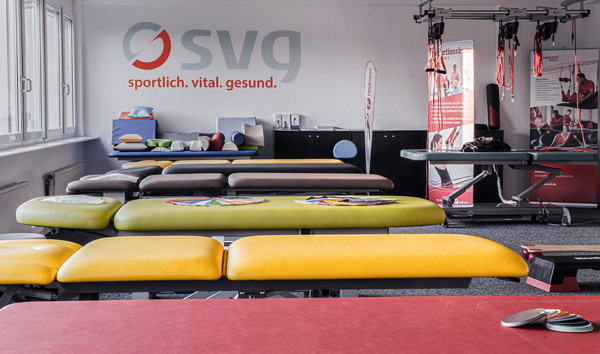 Behandlungsliegen in verschiedenen Farben stehen in einem Raum, an dessen Wand das SVG-Logo erkennbar ist